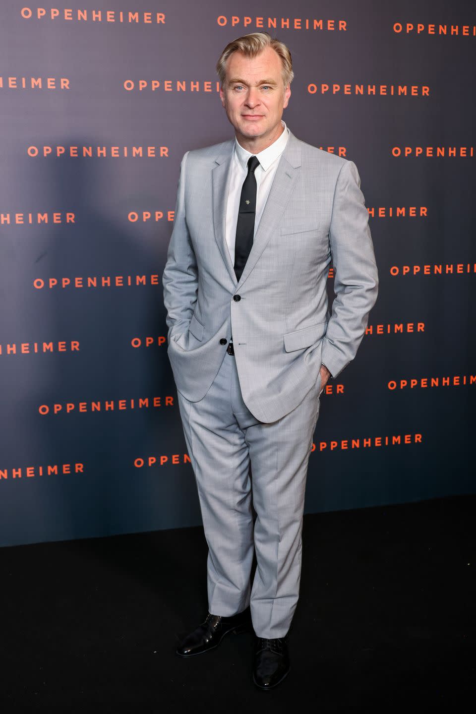 christopher nolan, a man stands with hands in trouser pockets, he wears a grey suit