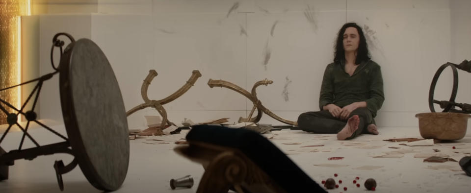 Tom Hiddleston as Loki sits barefoot in a disheveled room with broken furniture and scattered papers, looking somber