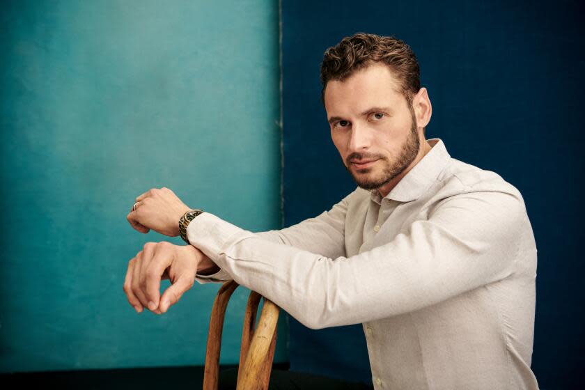 Adan Canto poses while resting his hands on a wooden chair, dressed in an off-white shirt