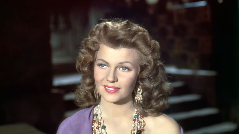rita hayworth smiles and looks left of the camera, she wears a mauve top with a purple scarf wrapped around her shoulders as well as a multistrand beaded necklace and large dangling earrings, she holds a card as she sits at a table with several other cards facing up