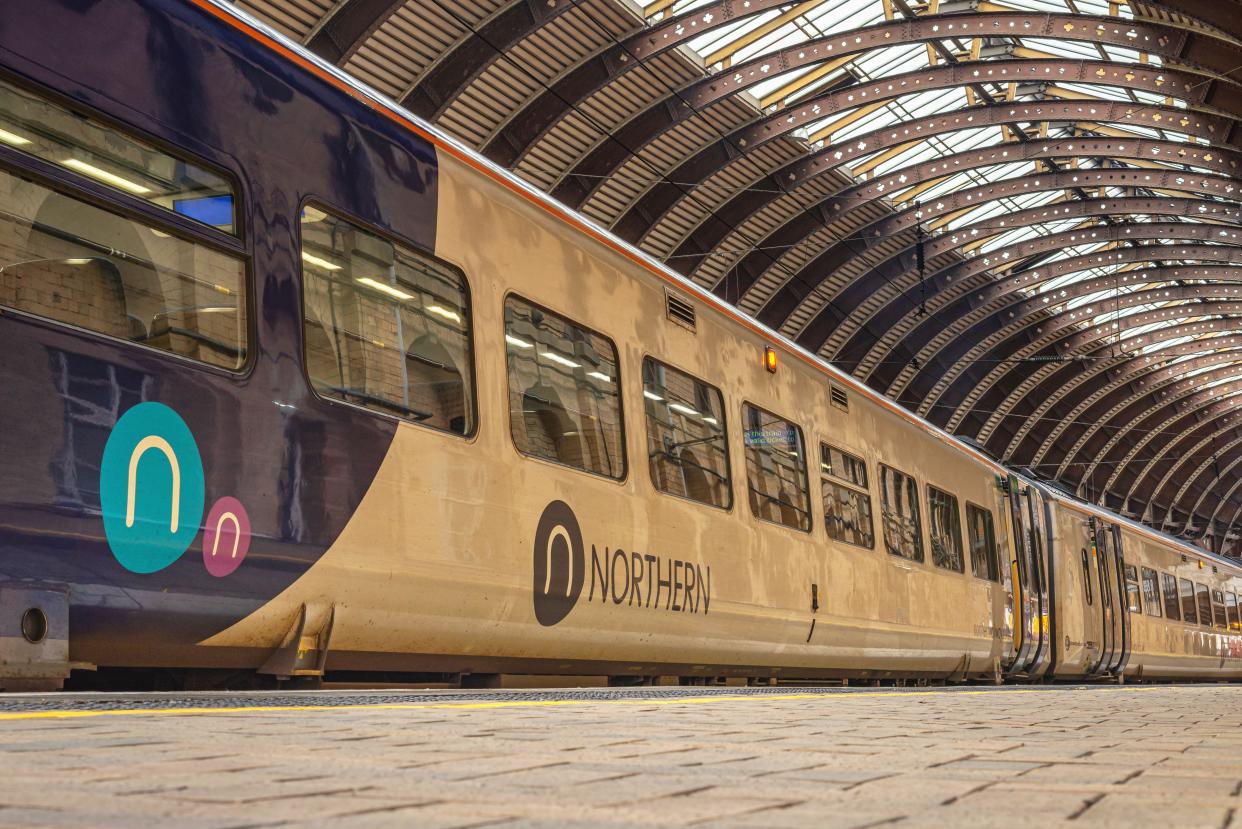 Two railway train carriages under a historic 19th Century metal canopy. A brick wall with arches is reflected in the widows of the carriage.