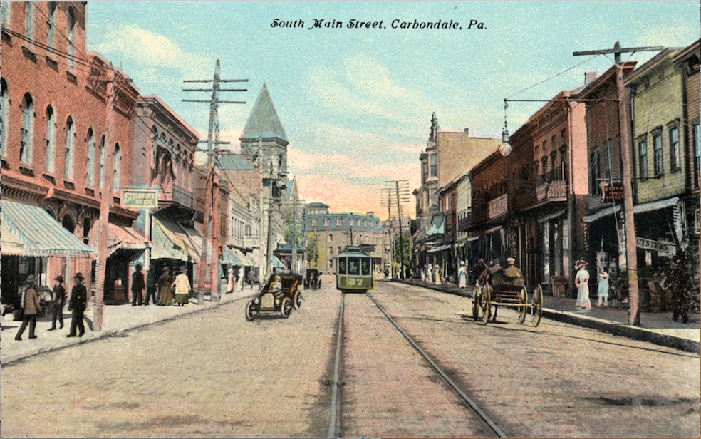 Bruning's Home Bakery, 28 S. Main St. Carbondale, Pa. is in one of these storefronts at left, thought to be the one where a woman in a light colored coat is walking under the awning, to the left of the passing car (which was drawn by an artist onto the original photograph to make the postcard appear more modern). City Hall is in the distance with the high belfry. Courtesy Susan White Pieroth, LackawannaGenWeb. Five Oaks Floral Shoppe is located at this address today.