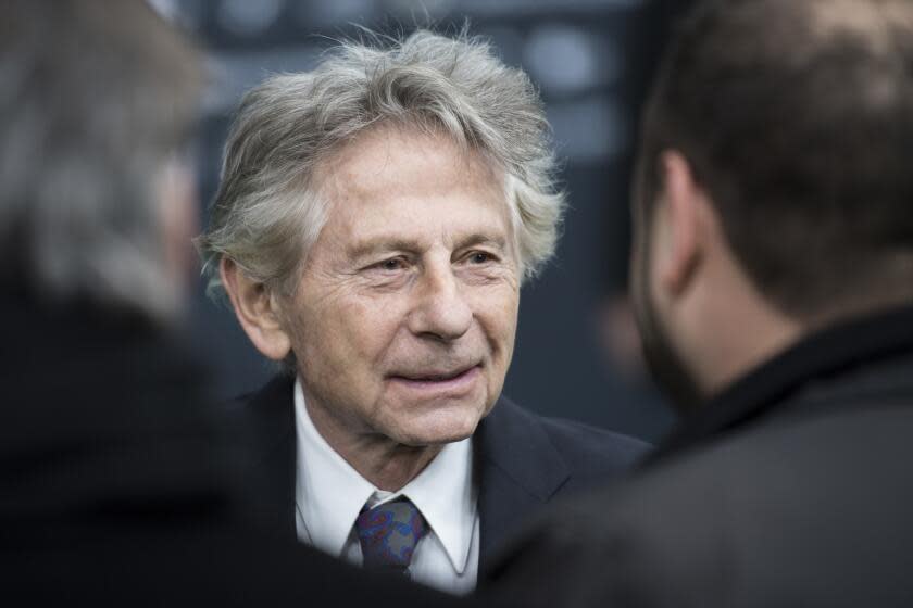 Roman Polanski wearing a black suit with a white undershirt and blue tie