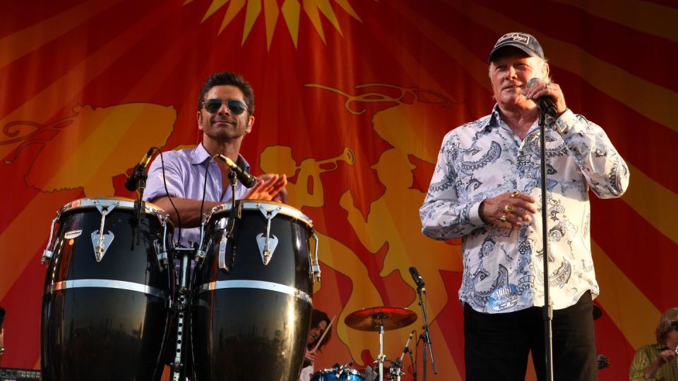 john stamos plays percussion on stage while wearing sunglasses and a purple short sleeved collared shirt, mike love of the beach boys stands to the right, love holds a microphone on a stand in one hand and wears black pants, a white paisley patterned shirt and a baseball cap