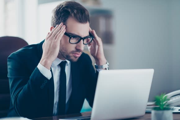 Bespectacled man in suit at laptop with annoyed expression, holding the sides of his head