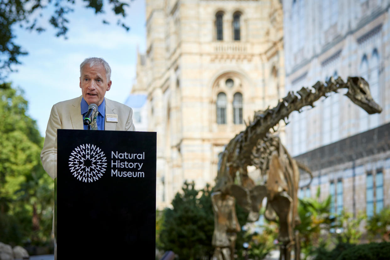 A man speaks at a lectern that features the Natural History Museum logo. There is a bronze dinosaur statue behind him and the museum building blurred in the background