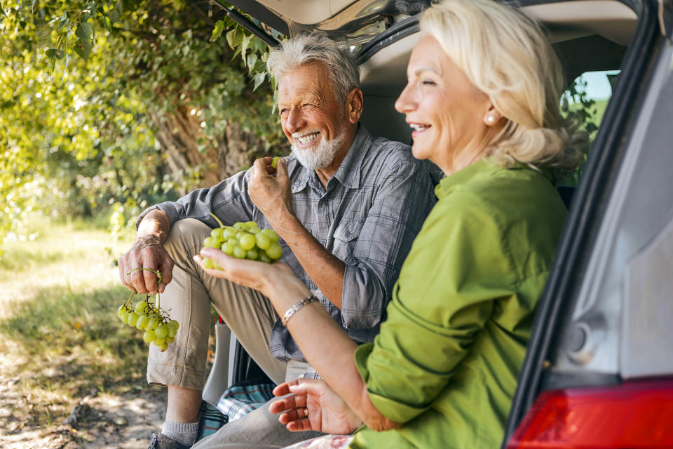 An older couple, both with greying hair, laugh and smile as they sit in the open trunk of a car and eat grapes together