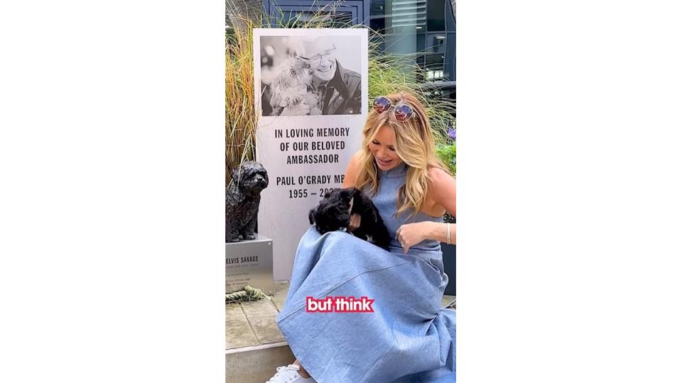 Amanda Holden with a black puppy in front of a memorial plaque for Paul O'Grady