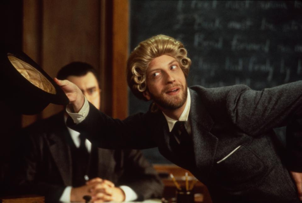 Person in period costume suit and curly wig, holding a hat and leaning forward with a playful expression. Looks like a classroom setting