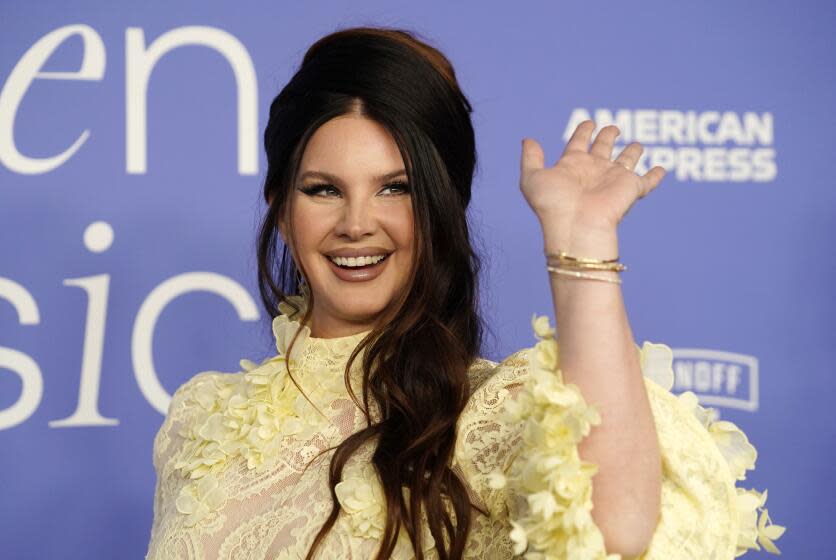 Lana del Rey is posing and smiling while waving her hand and is wearing a poofy yellow dress with embroidered designs