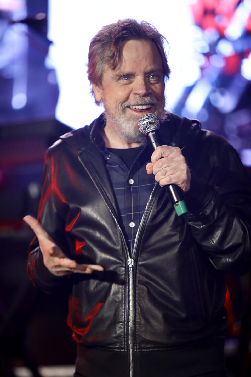 Mark talking animatedly into a microphone, wearing a black zipped up leather jacket over a dark plaid shirt