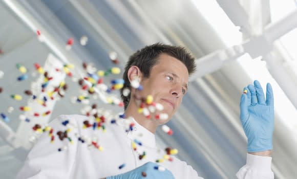 Man wearing white lab coat and blue gloves holding a pill, with several dozen pills scattered on a glass table.