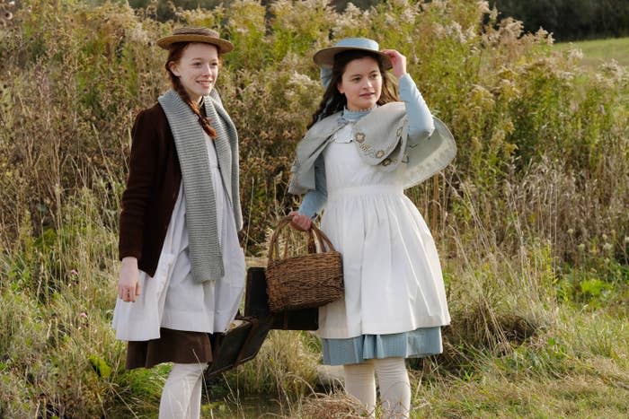 Two girls sand in a field wearing dresses, coats, and wide-brimmed hats. They each hold a picnic basket.