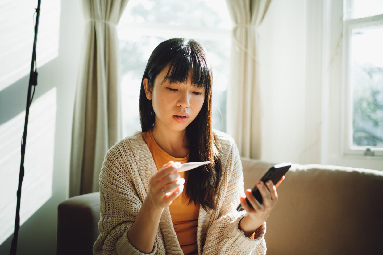 A woman sitting on a couch looks at an ovulation tracker while holding a smartphone.