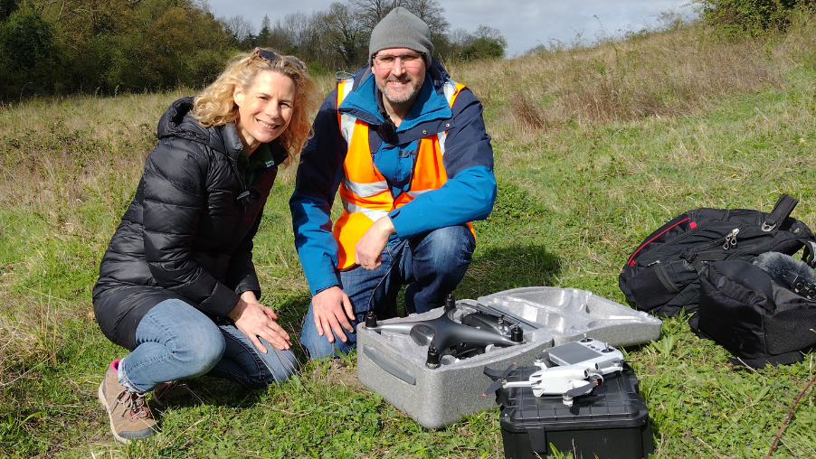 Emily Jeffery with drone pilot David Champion. They are in a field, both kneeling on the grass.