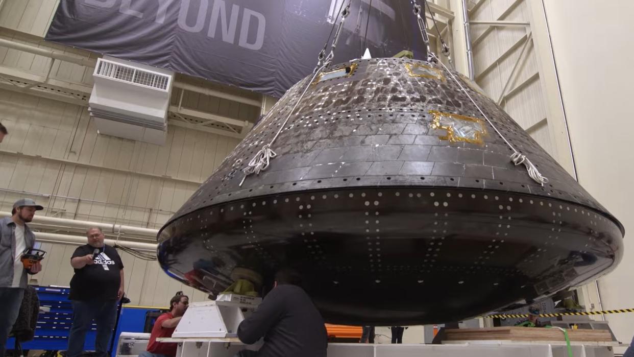  Close-up of a cone-shaped spacecraft in a warehouse, with people standing beside it. 