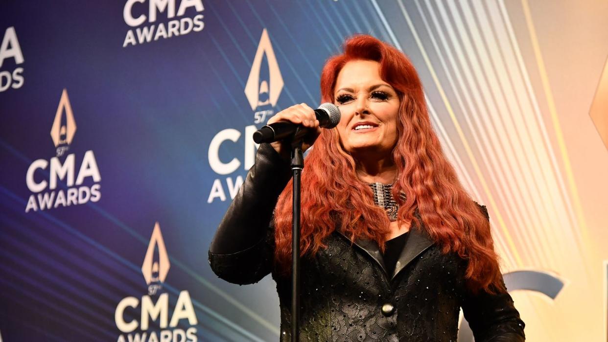 wynonna judd standing at a staged microphone and smiling for a photo