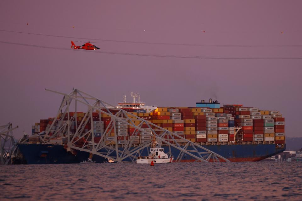 A collapsed bridge in front of a cargo ship, with a helicopter flying above.