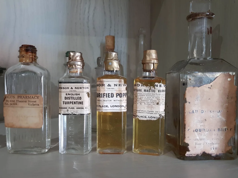 Five old bottles with various pharmaceutical labels are displayed on a shelf. Labels include: Aulis Pharmacy, Winsor & Newton Distilled Turpentine, Purified Poppy