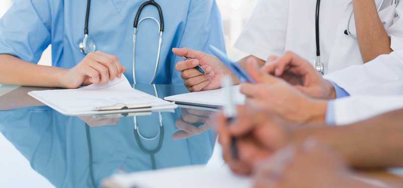 Several medical professionals sit in a group with stethoscopes and papers in hand.