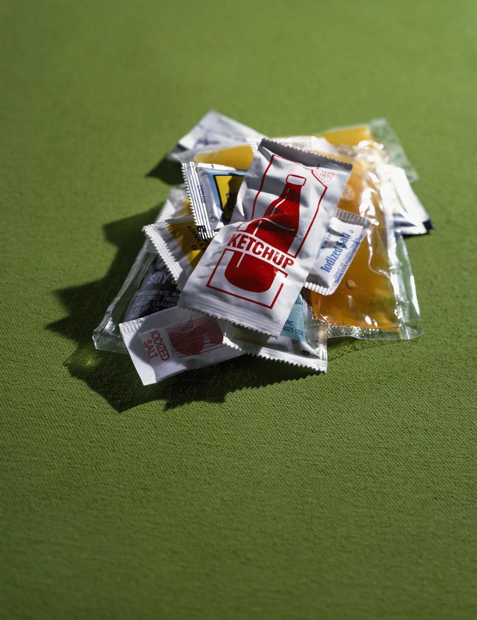 A small pile of assorted condiment packets, including ketchup, mustard, and soy sauce, placed on a green surface