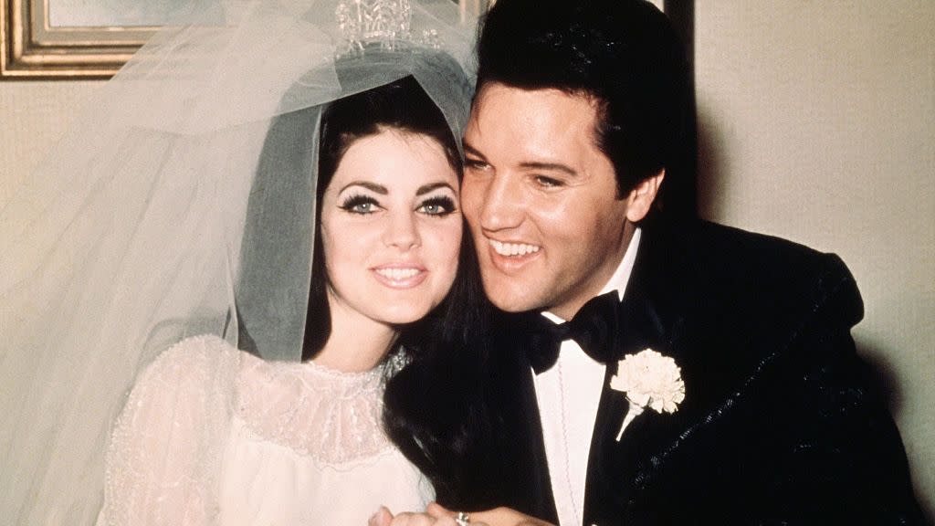 priscilla and elvis presley sit smiling and holding hands, she wears a wedding dress, veil and tiara, he wears a black suit with a bow tie and a carnation on his lapel