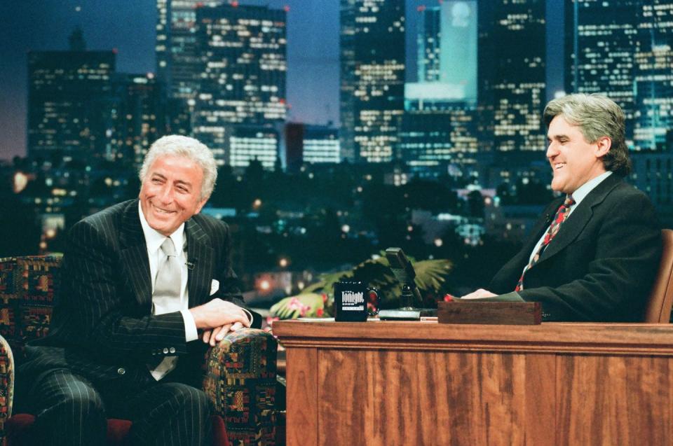 tony bennett smiles while leaning on the armrest of a couch, jay leno sits at a desk to the right and smiles at bennett, both men wear dark suits with ties