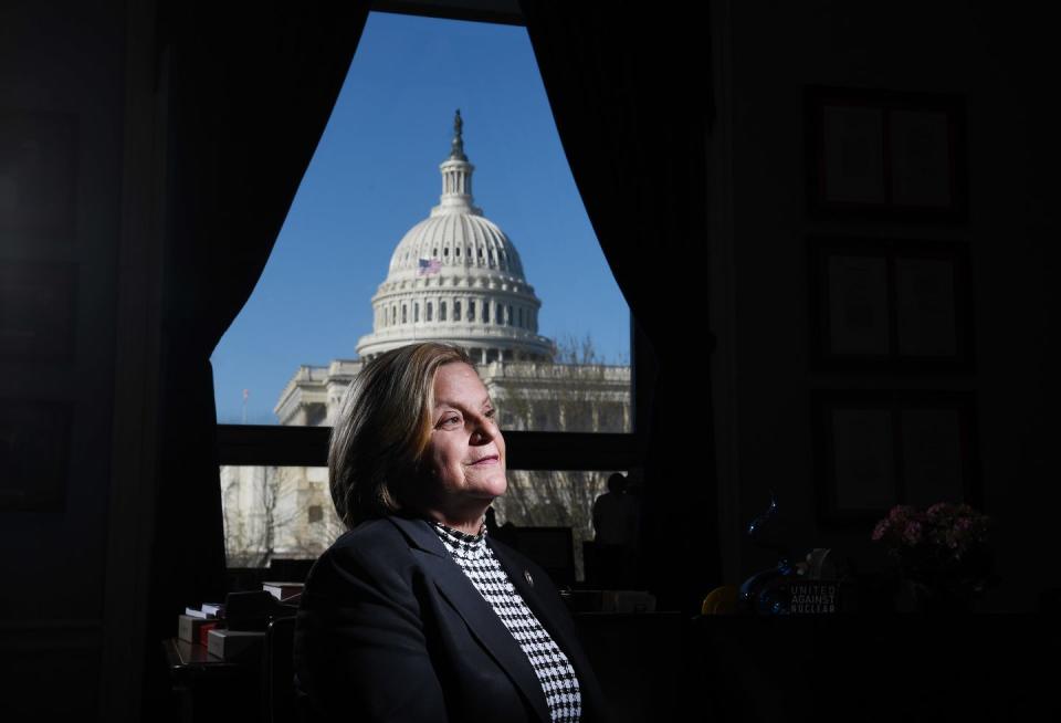 ileana ros lehtinen smiles and looks off into the distance as she sits in front of a window with curtains, outside is the dome of the captiol building, she wears a black suit jacket and black and white shirt