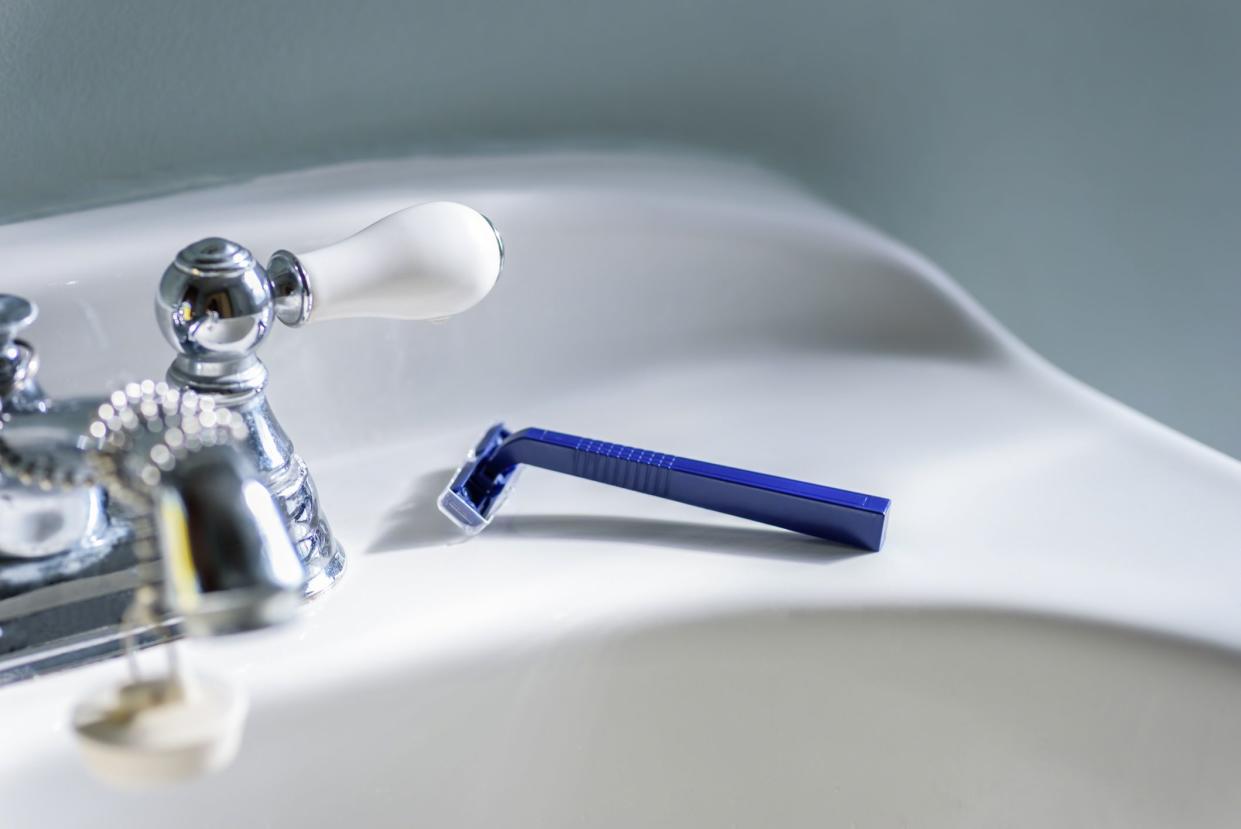 Selective focus of a blue disposable razor on ledge of a white sink in bathroom, near to faucet, with a seafoam colored wall in the background