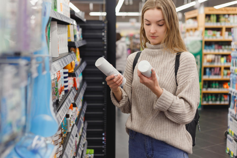 young woman picking bottles with shampoo and conditioner from shelf in cosmetics store�