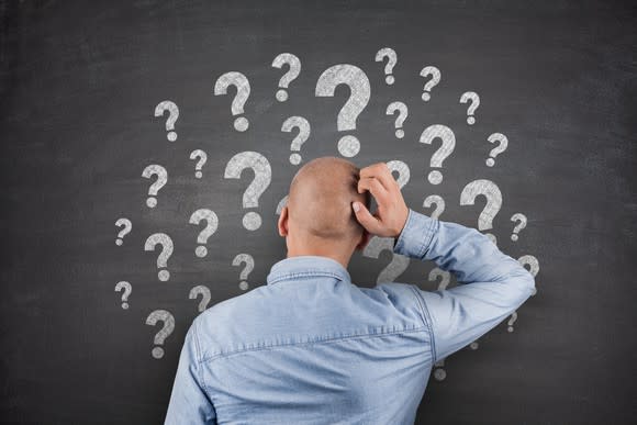 Man scratching head in front of chalkboard with many question marks drawn on it