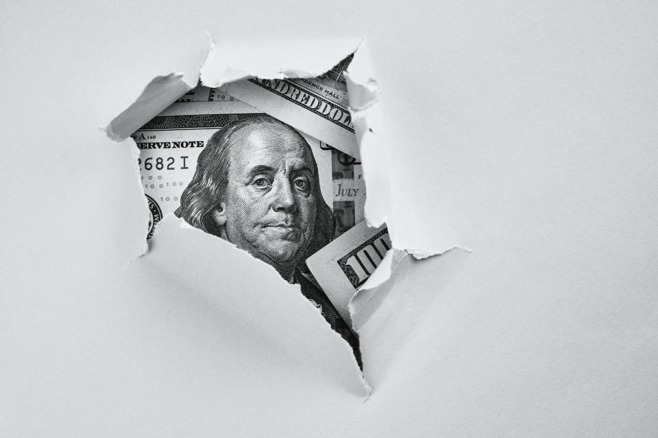 A ripped hole in a white piece of paper revealing a $100 bill hidden underneath.