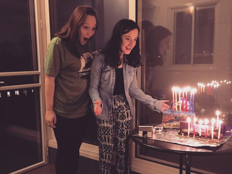 Two women look at a menorah lit with Hanukkah candles with happy expressions.