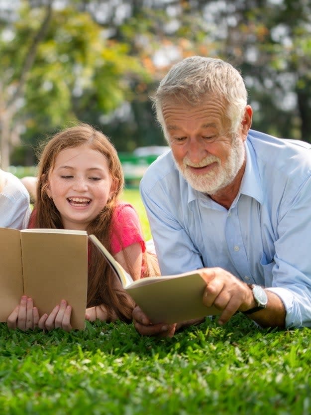 An elderly man and a young girl lie on the grass and read books together, smiling