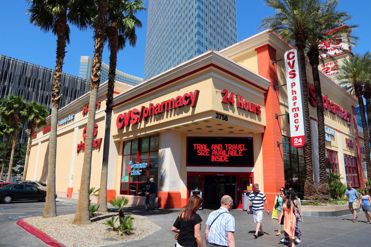 Front exterior of a CVS Pharmacy store in Las Vegas with people walking by on a large sidewalk, surrounded by palm trees with casino buildings and a blue sky in the background