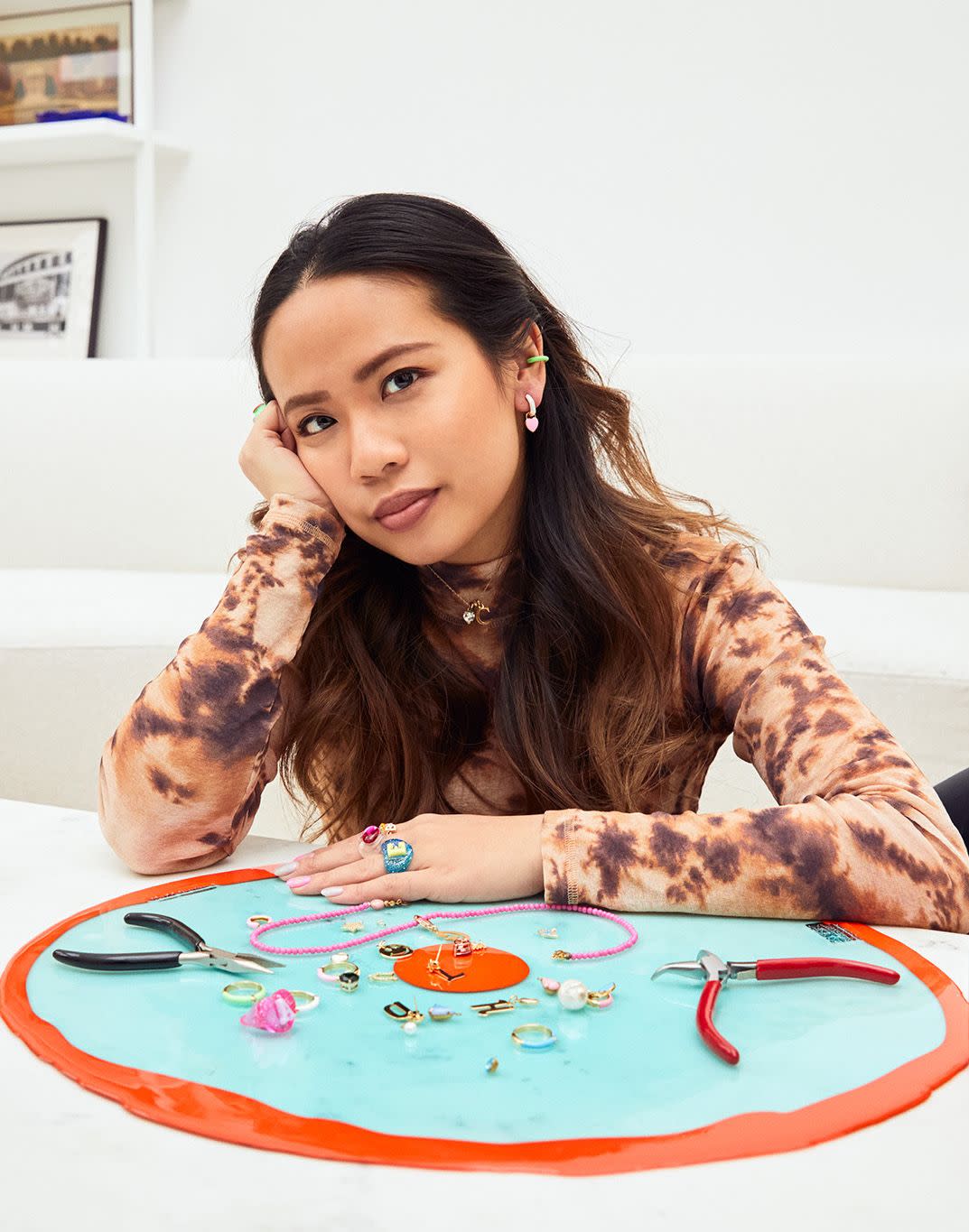 designer clare ngai sits in front of a turquoise and orange tray of colorful jewelry and pliers