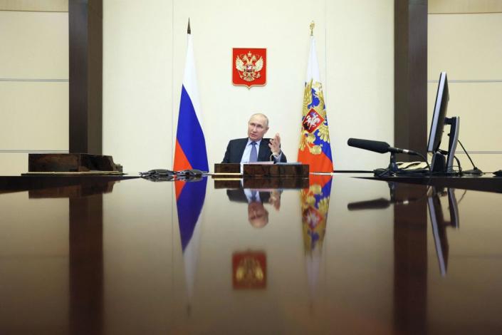 Vladimir Putin seen from the end of an extremely long, shiny table, with two flags beside him and a gold, double-headed eagle on a crimson background on the wall above him. 