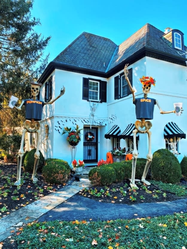 Two skeletons wearing "Pumpkin" and "Spice" T-shirts and holding large coffee cups in front of a house