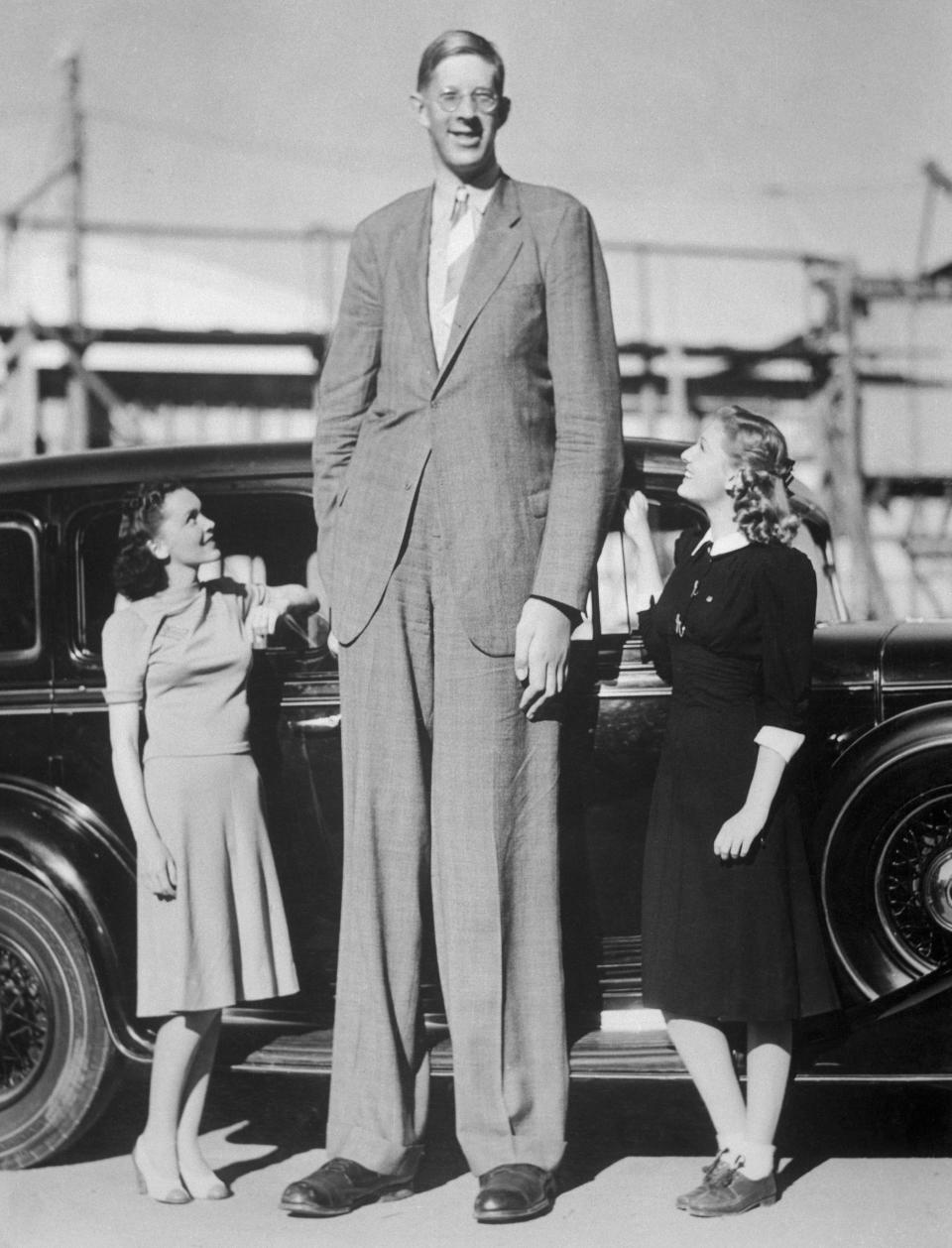 He's wearing a suit and standing in front of a car next to two smiling women, one of whom comes to his hip and another to his waist