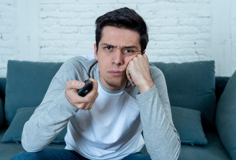 Lifestyle portrait of young bored man on couch with remote control zapping for movie or live sport. Looking disinterested drinking beer. Sedentary and mass social media or Television addiction.