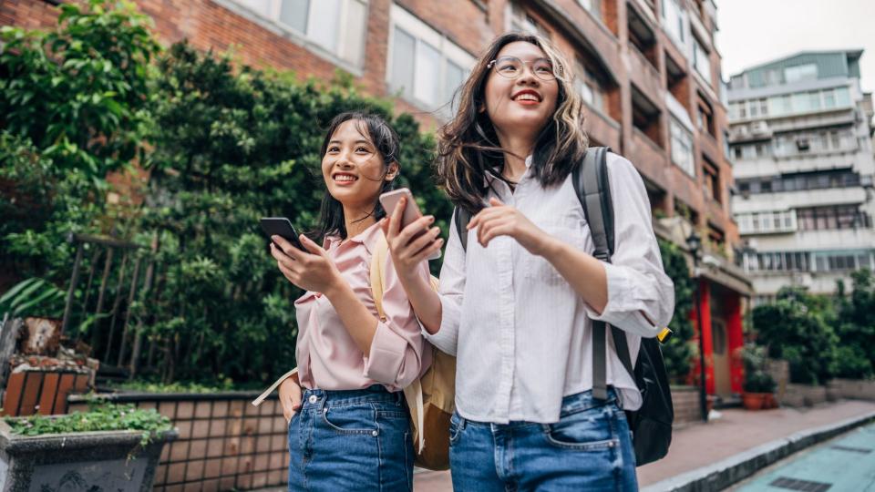 Two women, young women students together in city on the street, using smart phones, looking for apartment.