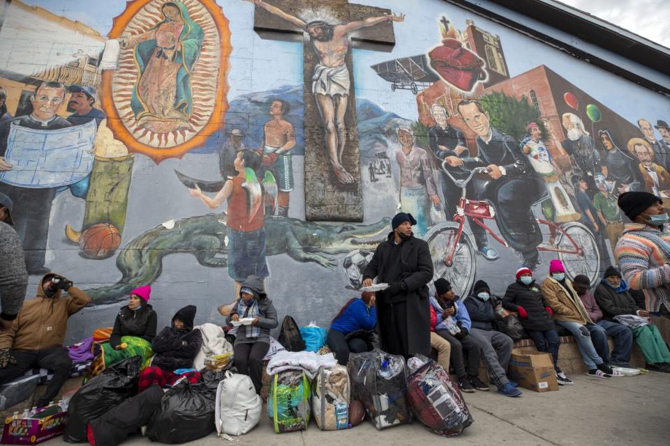 People with baggage sit on the sidewalk outside beneath a mural.