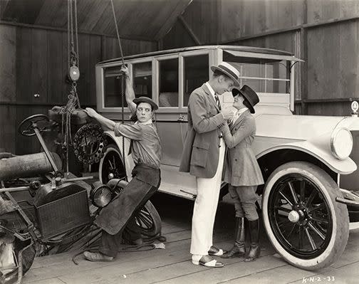 Buster Keaton hoists a Model T engine from a wrecked car as Virginia Fox flirts with an unidentified actor in a scene still for the 1922 comedy short The Blacksmith.