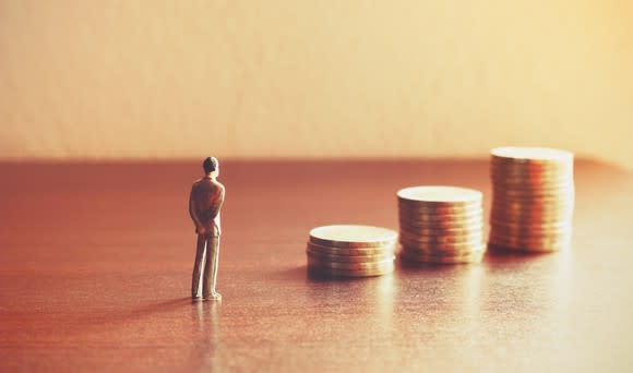 A tiny man stands in front of stacks of coins.