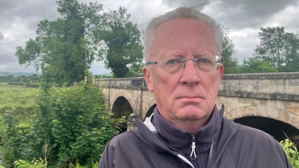 Eamon Mullan from the shoulders up looking at the camera and standing in front of River Roe bridge