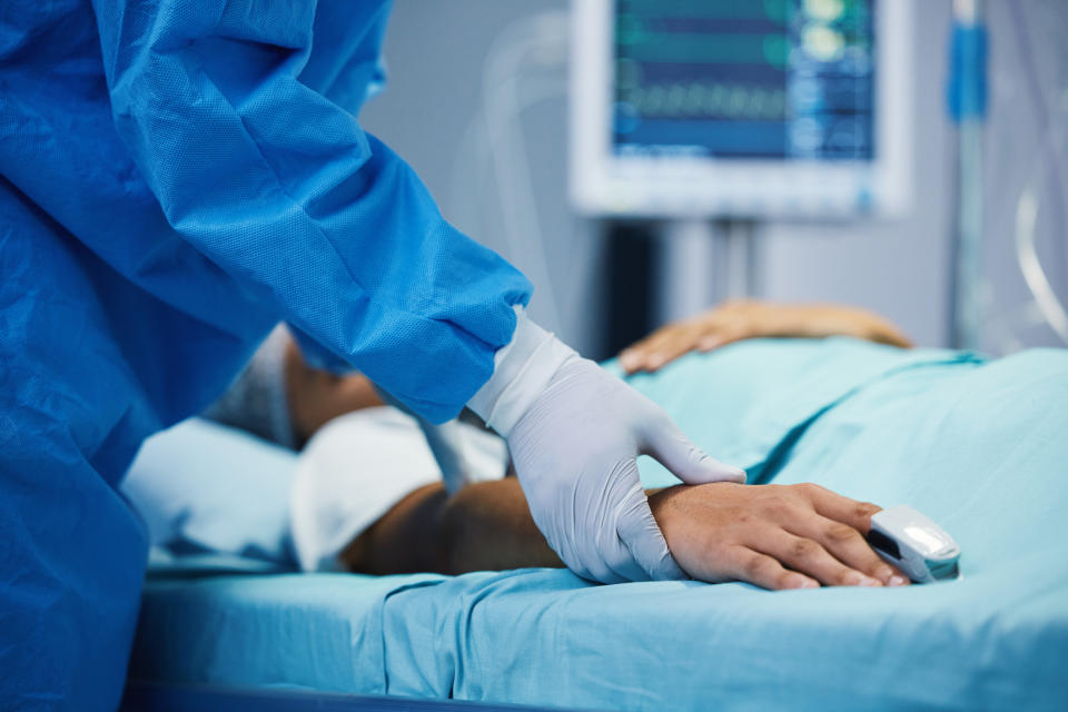 A healthcare worker wearing protective gloves and a gown checks the pulse of a patient's hand in a hospital bed. Medical equipment is visible in the background