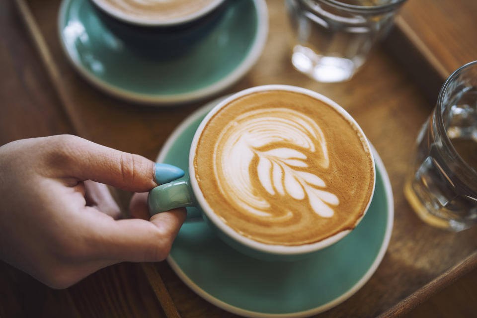 A hand holds a cup of coffee with a beautiful leaf design in the foam, next to a glass of water on a wooden tray