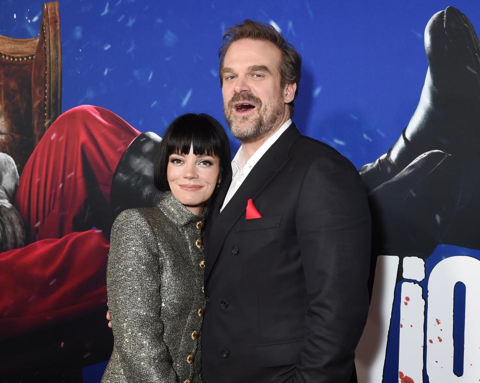 David Harbour and Lily Allen stand together, smiling, at a movie premiere. David wears a dark suit with a bright pocket square, and Lily wears a stylish buttoned coat