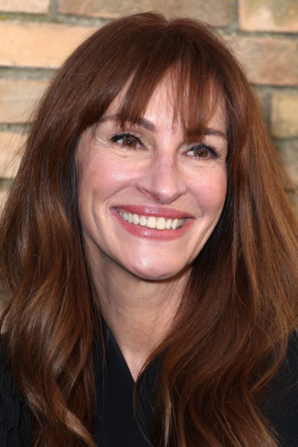 Smiling woman with long brown hair and bangs, standing in front of a brick wall. Names unknown