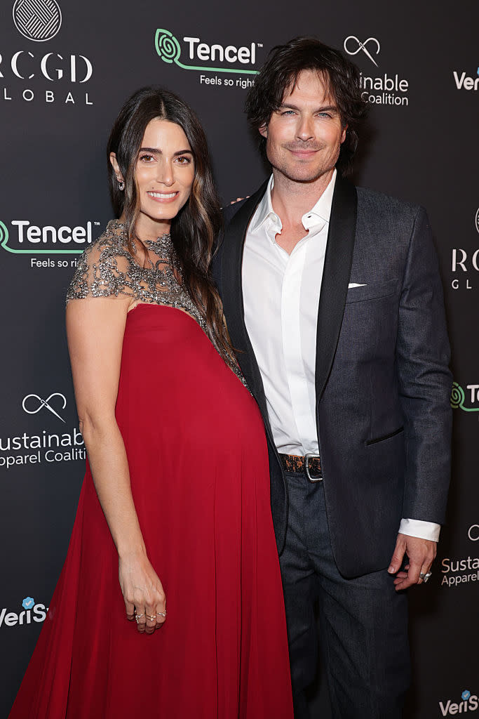 Nikki Reed in a flowing, embellished dress and Ian Somerhalder in a suit, posing together at an event. Nikki is visibly pregnant